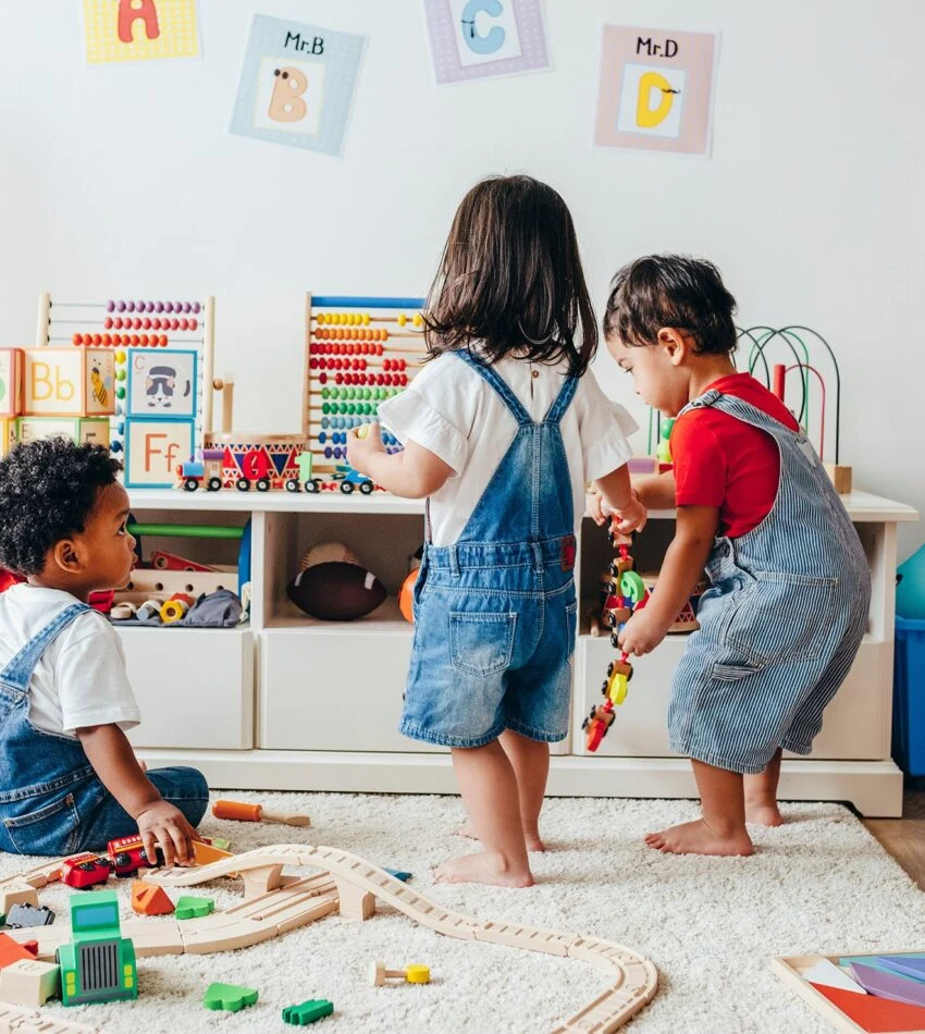 Two toddlers in denim overalls playing with toys and a wooden train in a bright playroom